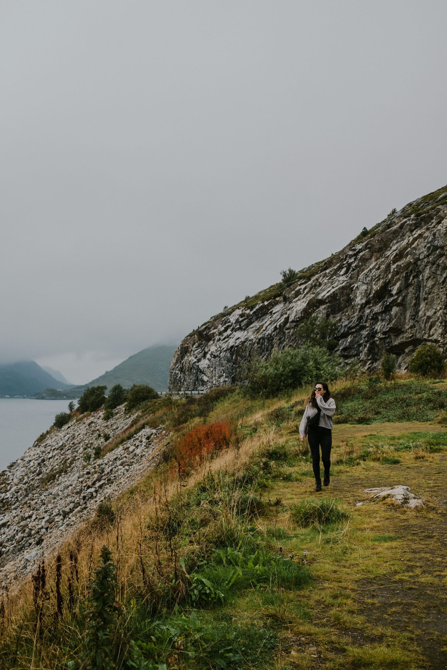 Hiker on coastal trail along dramatic rocky cliffs beside misty fjord.