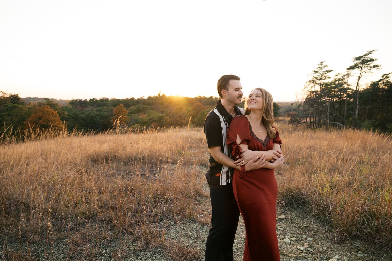A romantic couple shares a laugh while embracing on a trail through tall grass at sunset.