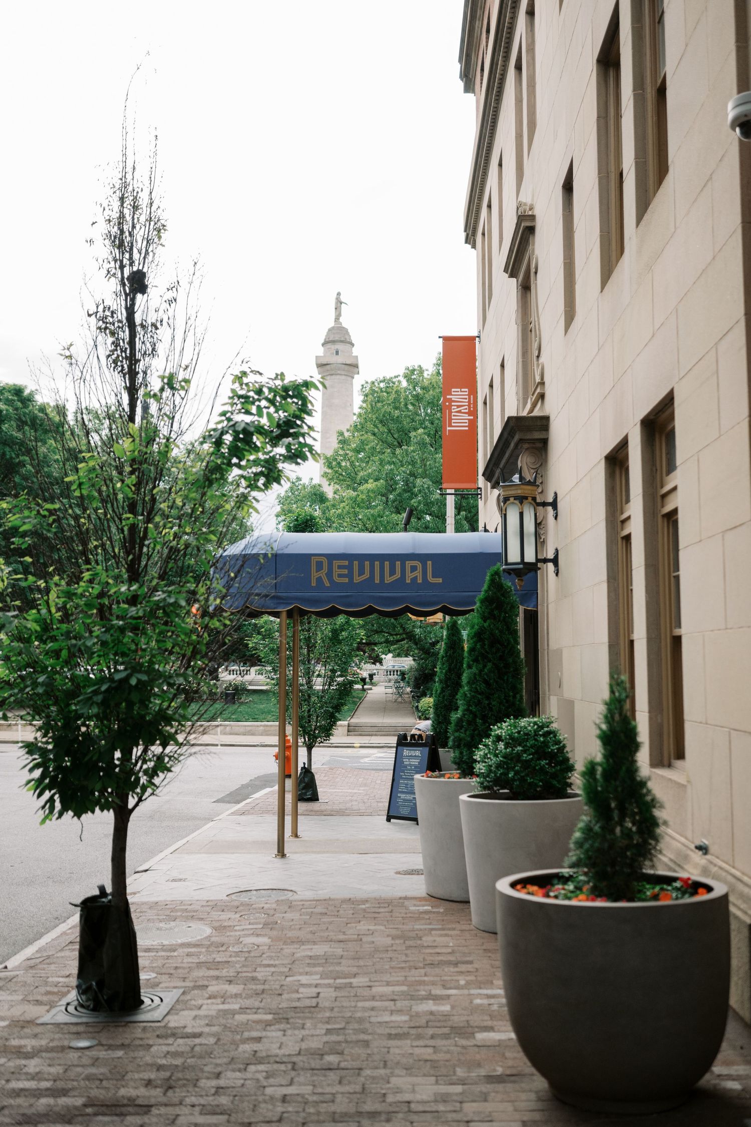 Urban sidewalk with potted plants and trees leading to a city building with a blue 'Revival' sign.