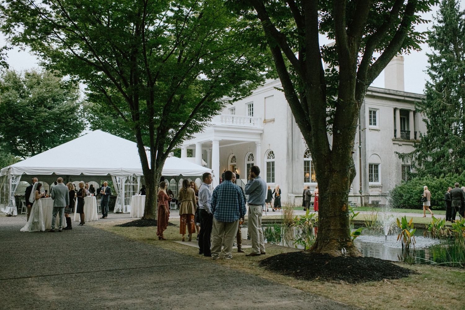 Wedding reception venue with white tent and guests mingling on tree-lined path near elegant house.