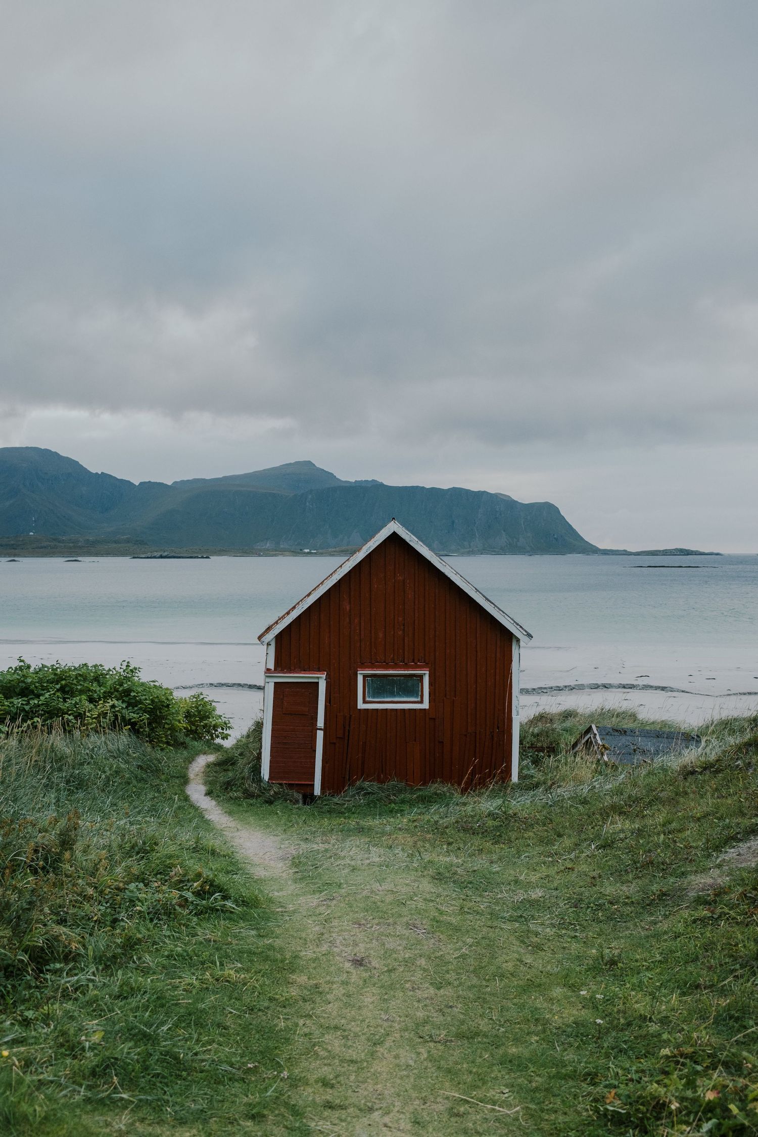Red wooden cabin on grassy path overlooking serene fjord and mountain landscape.