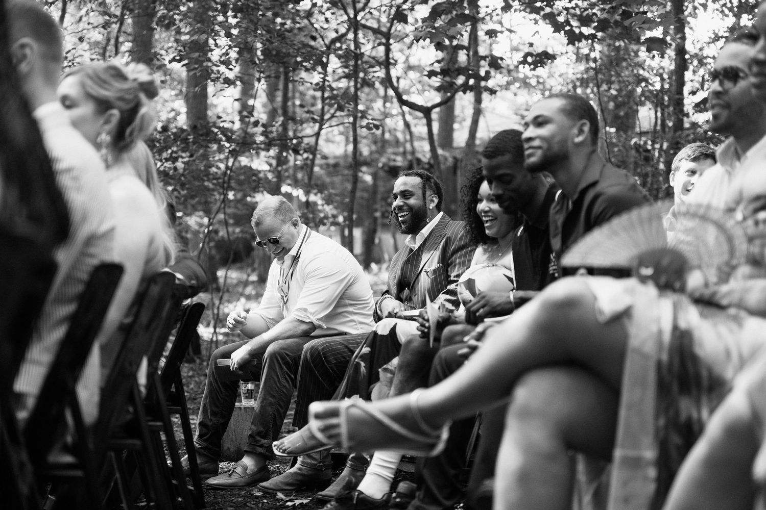 Wedding guests seated on wooden chairs watch the ceremony in a wooded setting.