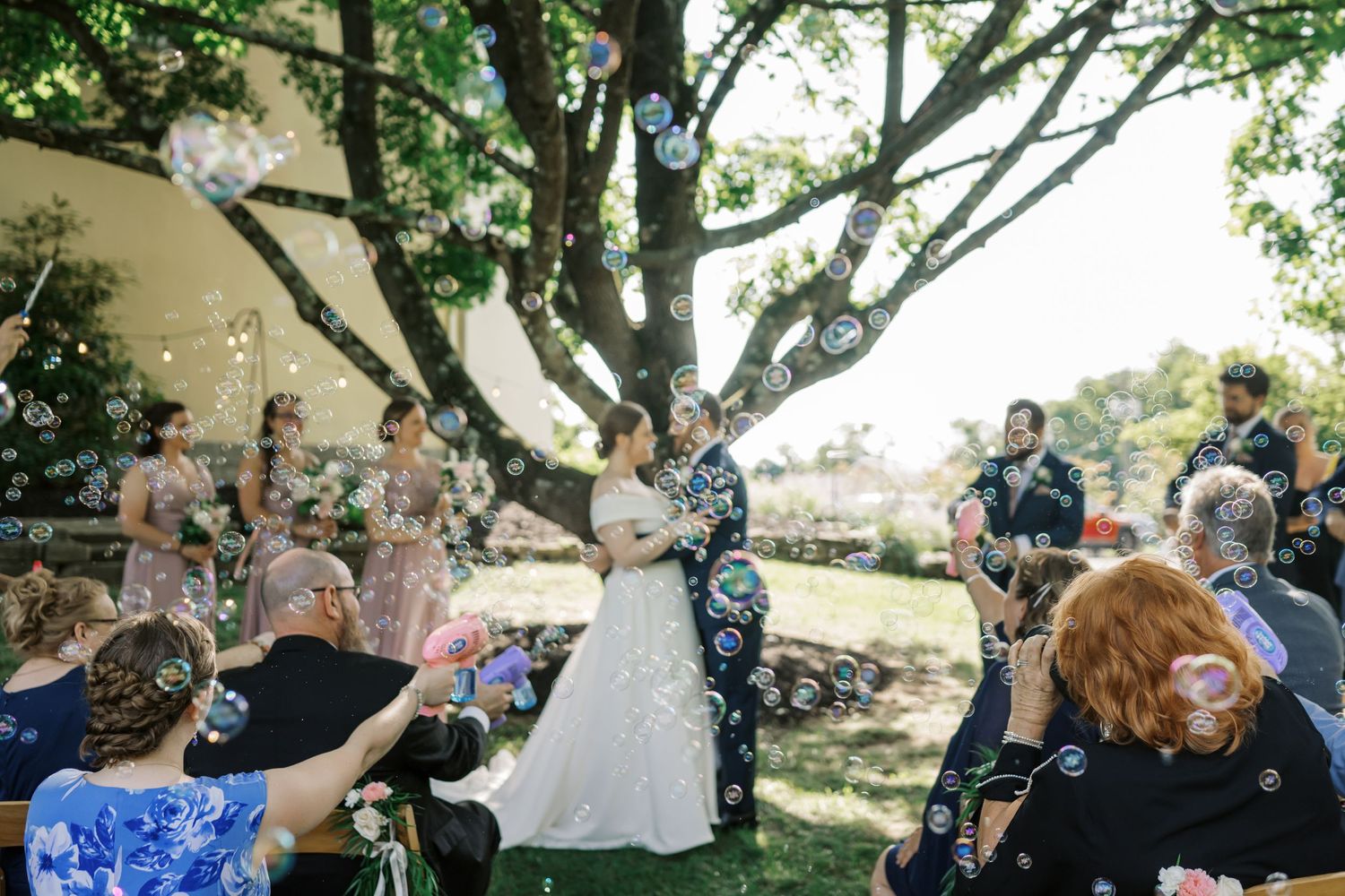 Wedding guests blow bubbles at a couple during an outdoor garden ceremony.
