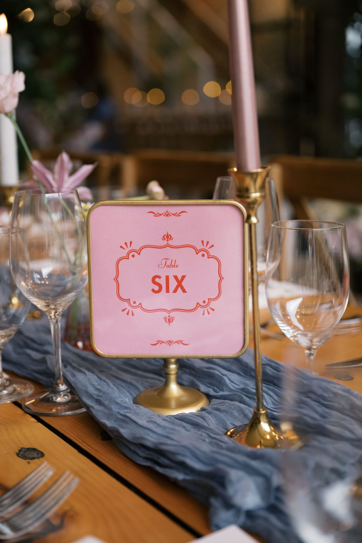 Pink table number six sign on gold stand with wine glasses and blue table runner.