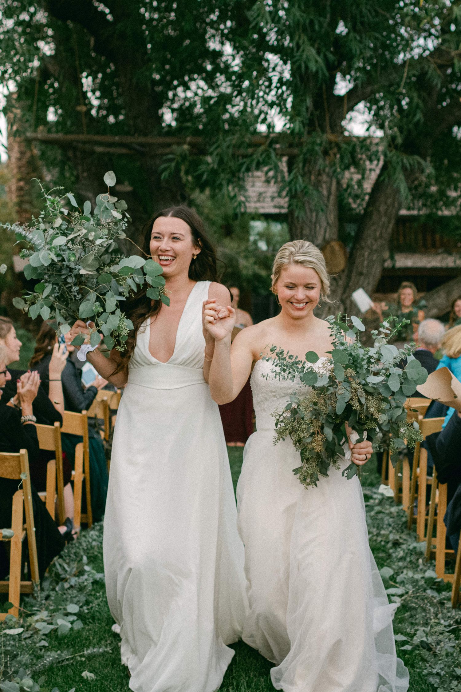 Two people in white dresses celebrate with bouquets while walking down the aisle at an outdoor ceremony.