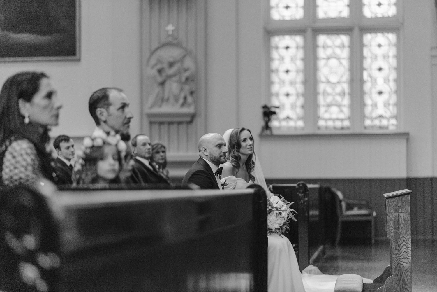 Black and white photo of wedding ceremony attendees seated in wooden church pews.