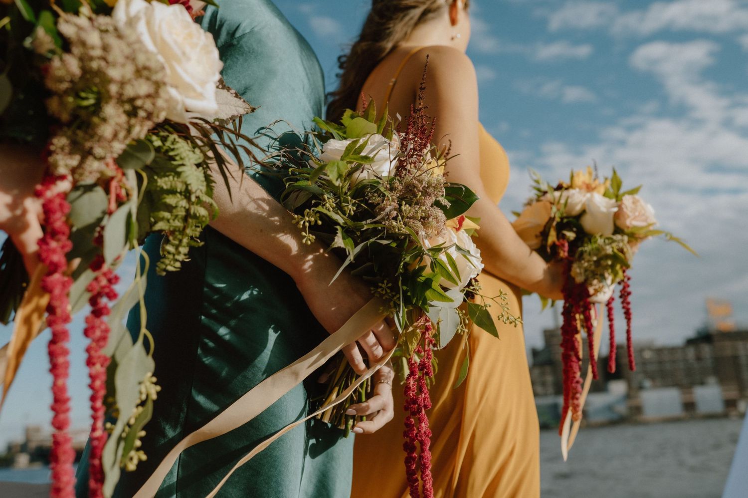 Wedding party members hold elegant bouquets with cascading flowers and ribbons against a blue sky.