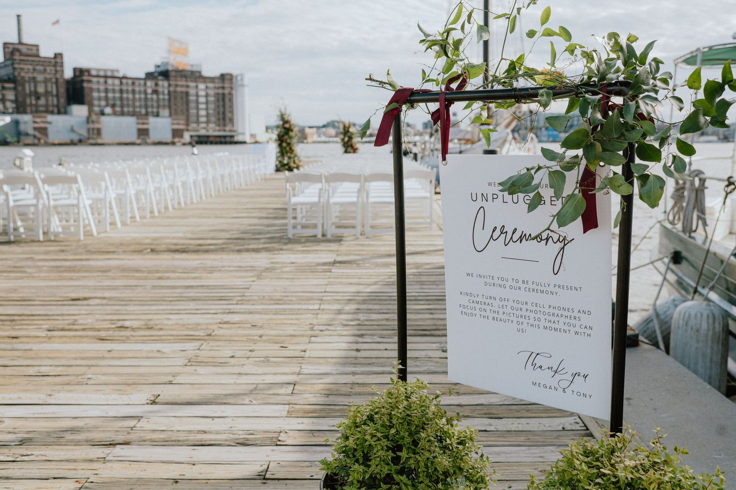 Waterfront wedding ceremony setup with white chairs and floral decorated welcome sign on dock.