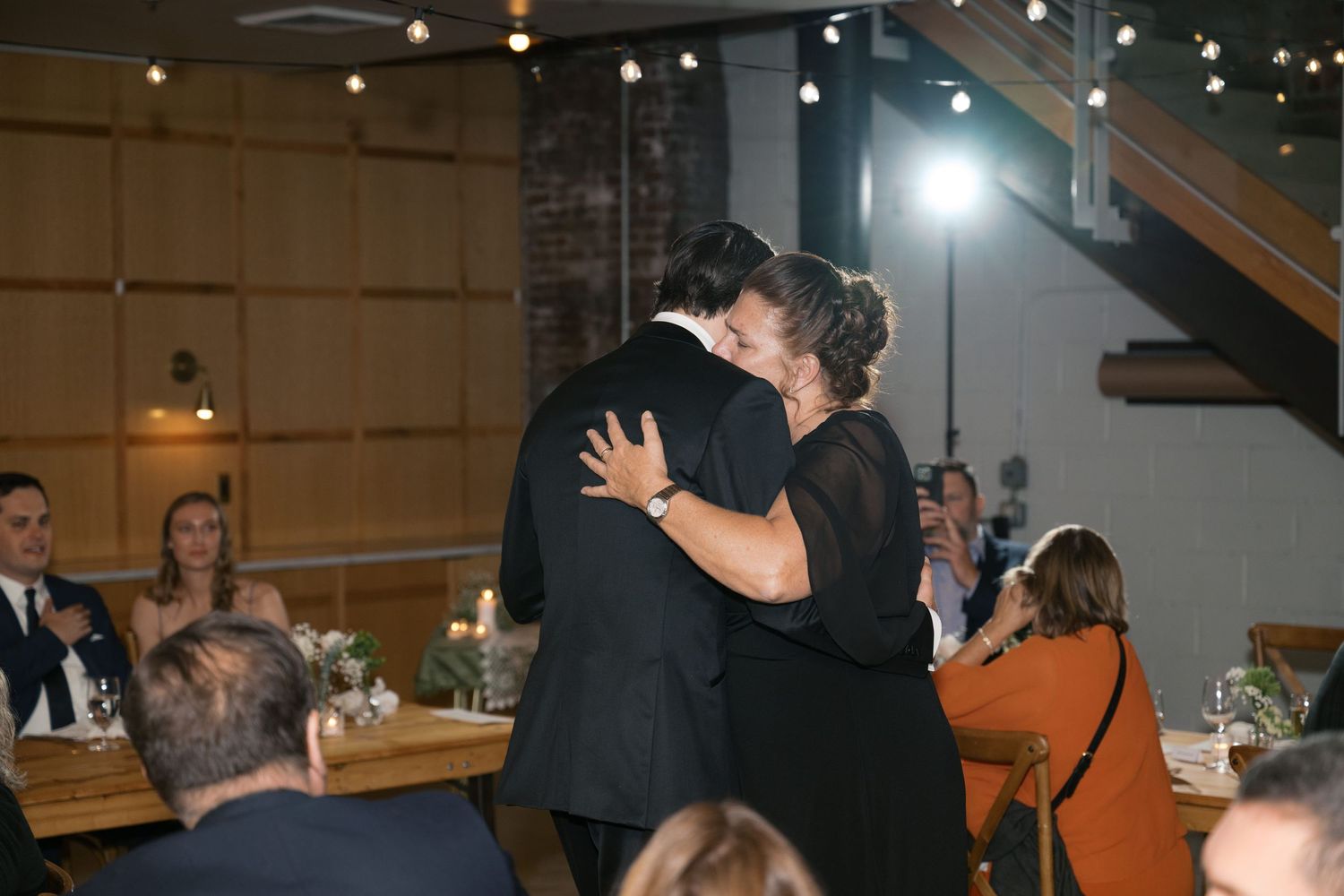 A couple dances together at a reception venue with warm lighting and guests seated at tables in the background.
