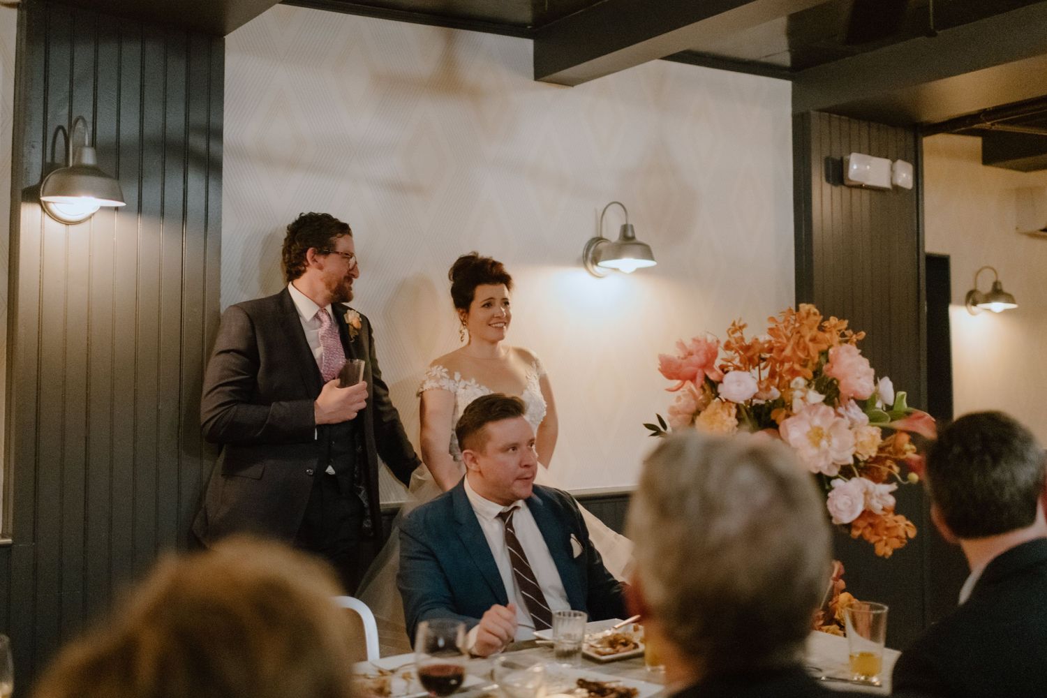 Wedding reception scene with elegant floral centerpieces and pendant lighting.