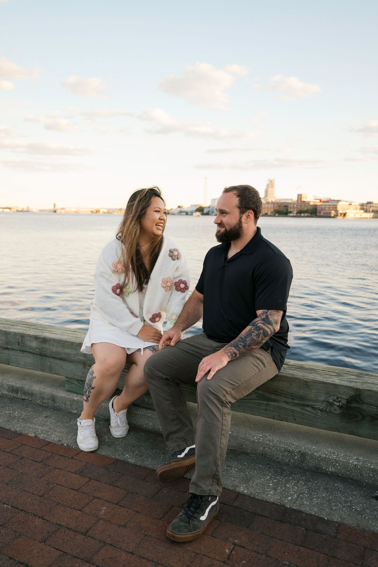 A couple sits together on a concrete wall by the waterfront at sunset, sharing a relaxed moment overlooking the ocean.