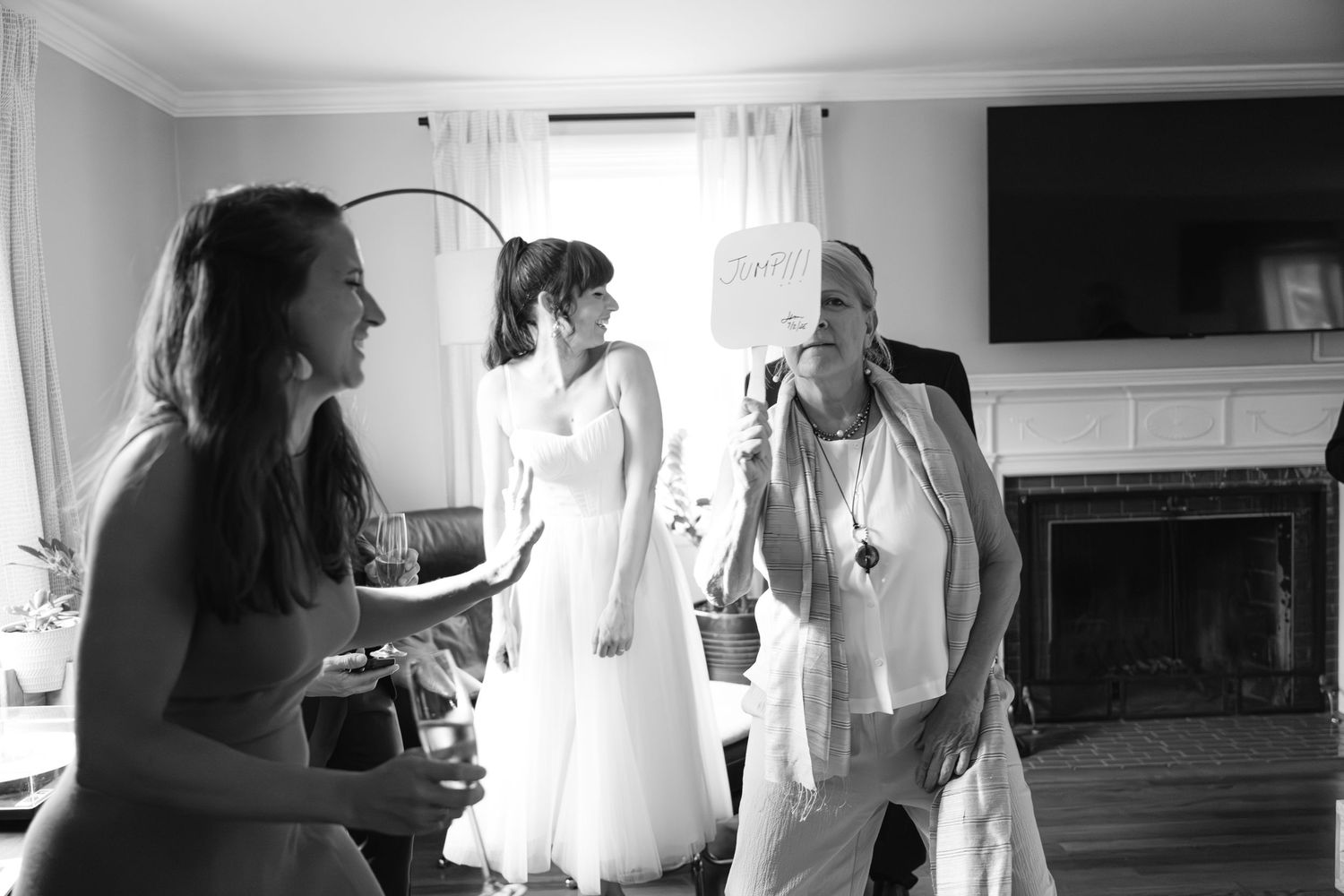 A candid black and white scene showing joyful interactions during wedding preparations in a bright room with a fireplace.