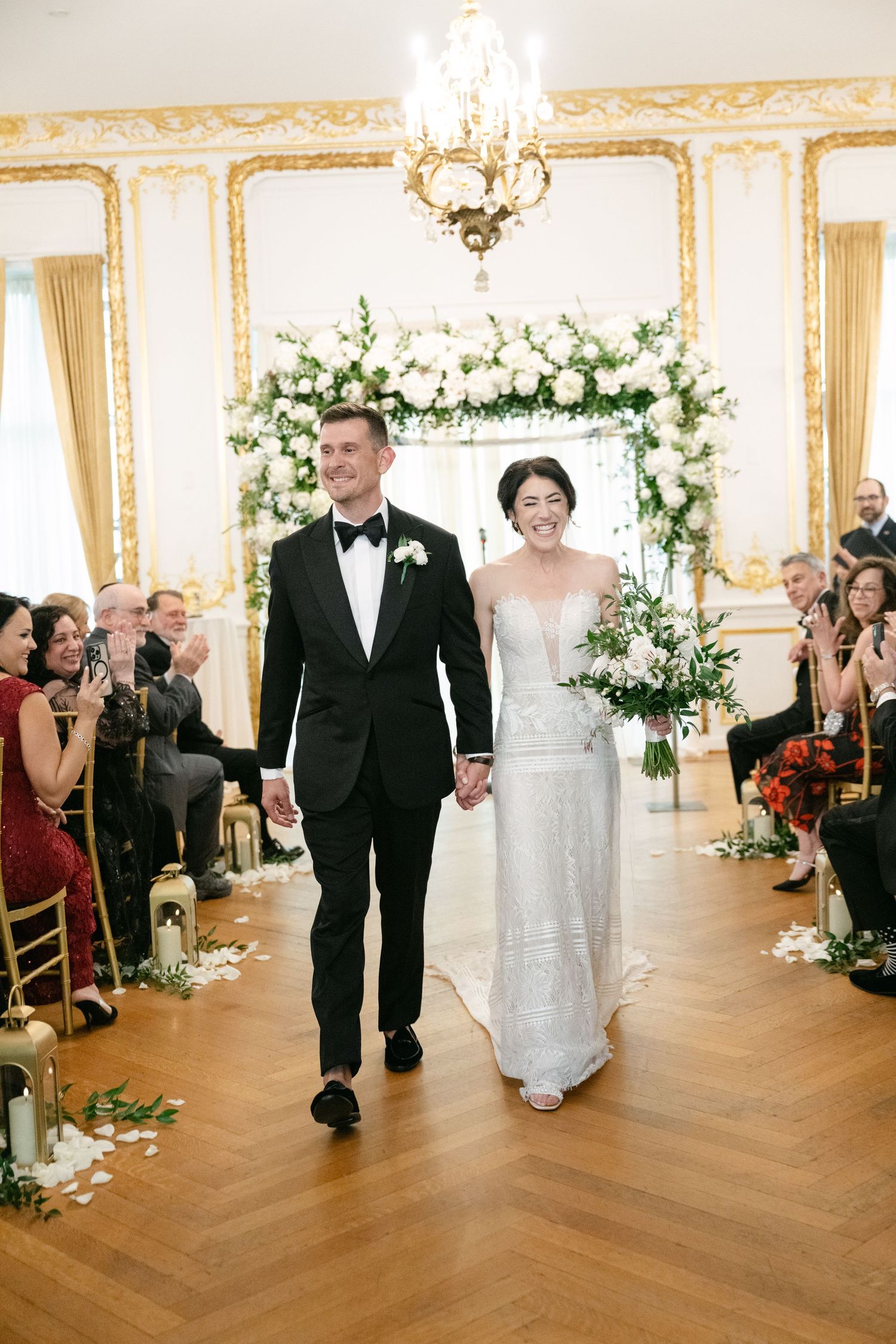 Wedding ceremony in an elegant ballroom under a floral arch with guests seated on gold chairs.