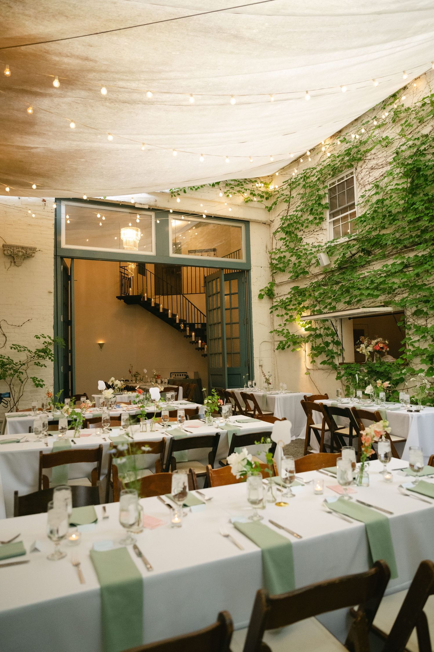 An elegant indoor reception venue with long white dining tables set up under a fabric canopy with string lights and greenery.