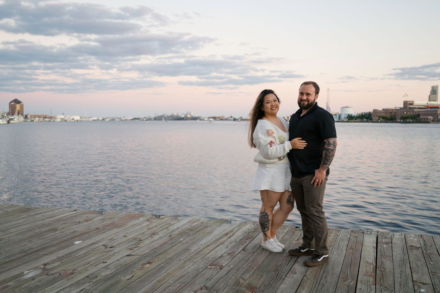 Two people share a romantic moment on a waterfront pier at sunset with a calm harbor and cityscape in the background.