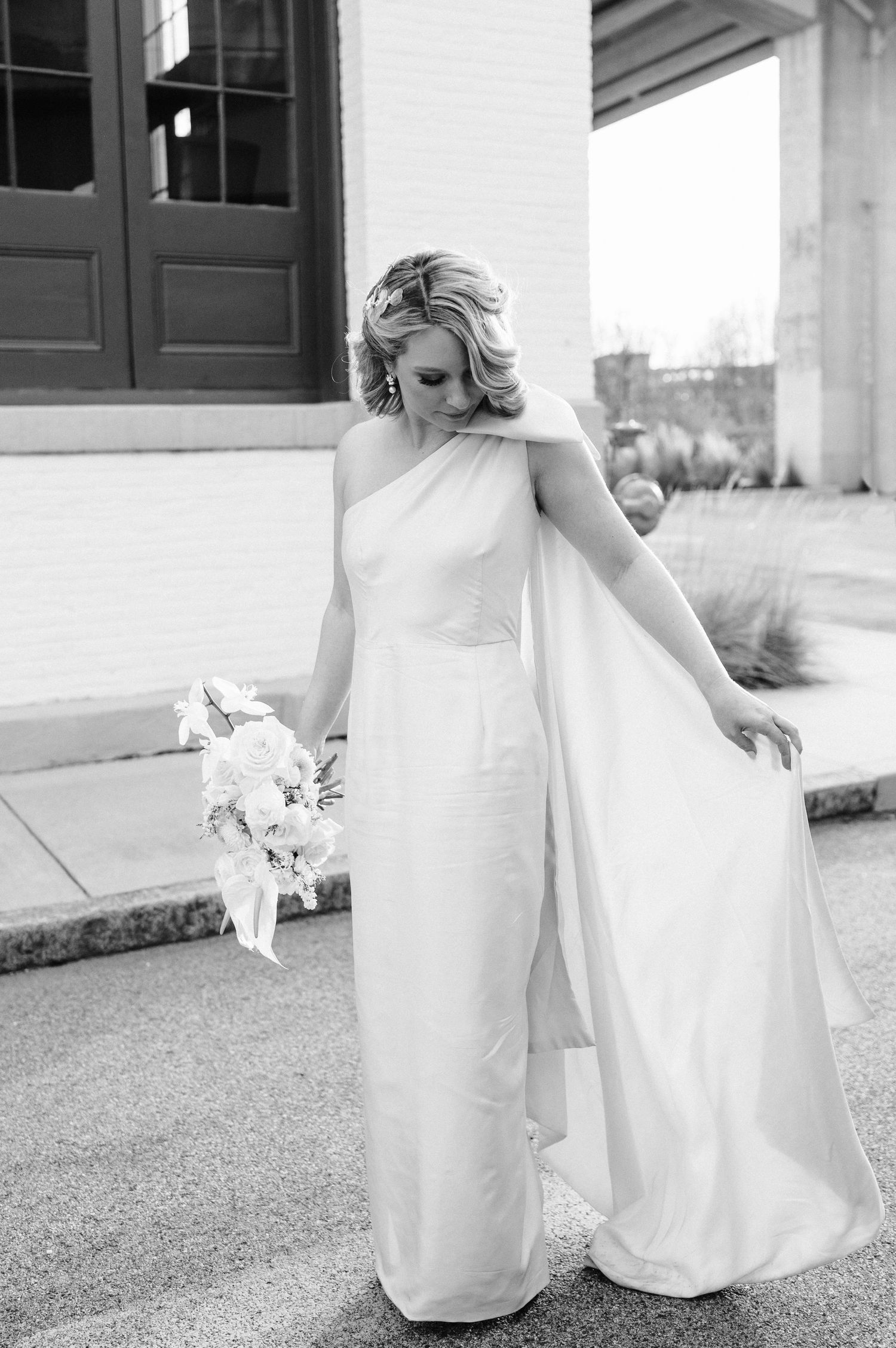 Black and white portrait of bride in elegant one-shoulder wedding dress holding bouquet.