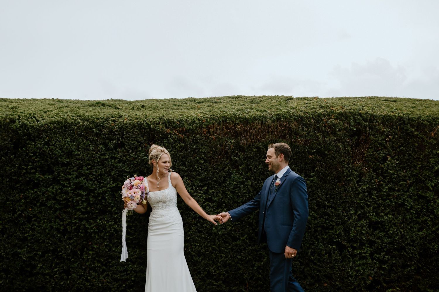 A couple poses for a wedding portrait against a tall manicured hedge wall.
