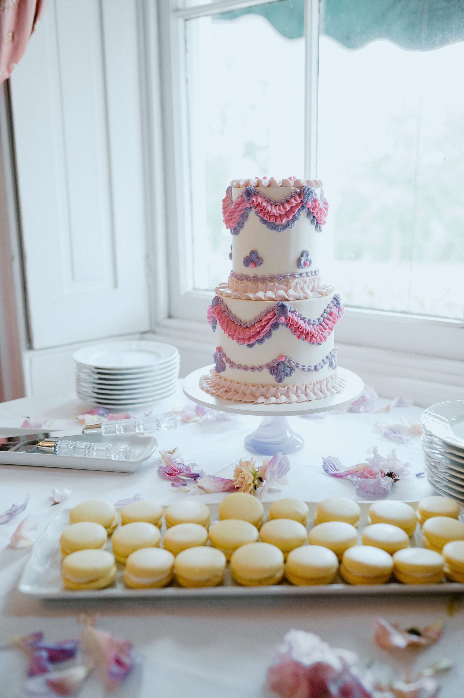 Two-tier white wedding cake with pink piping and yellow macarons displayed on dessert table.
