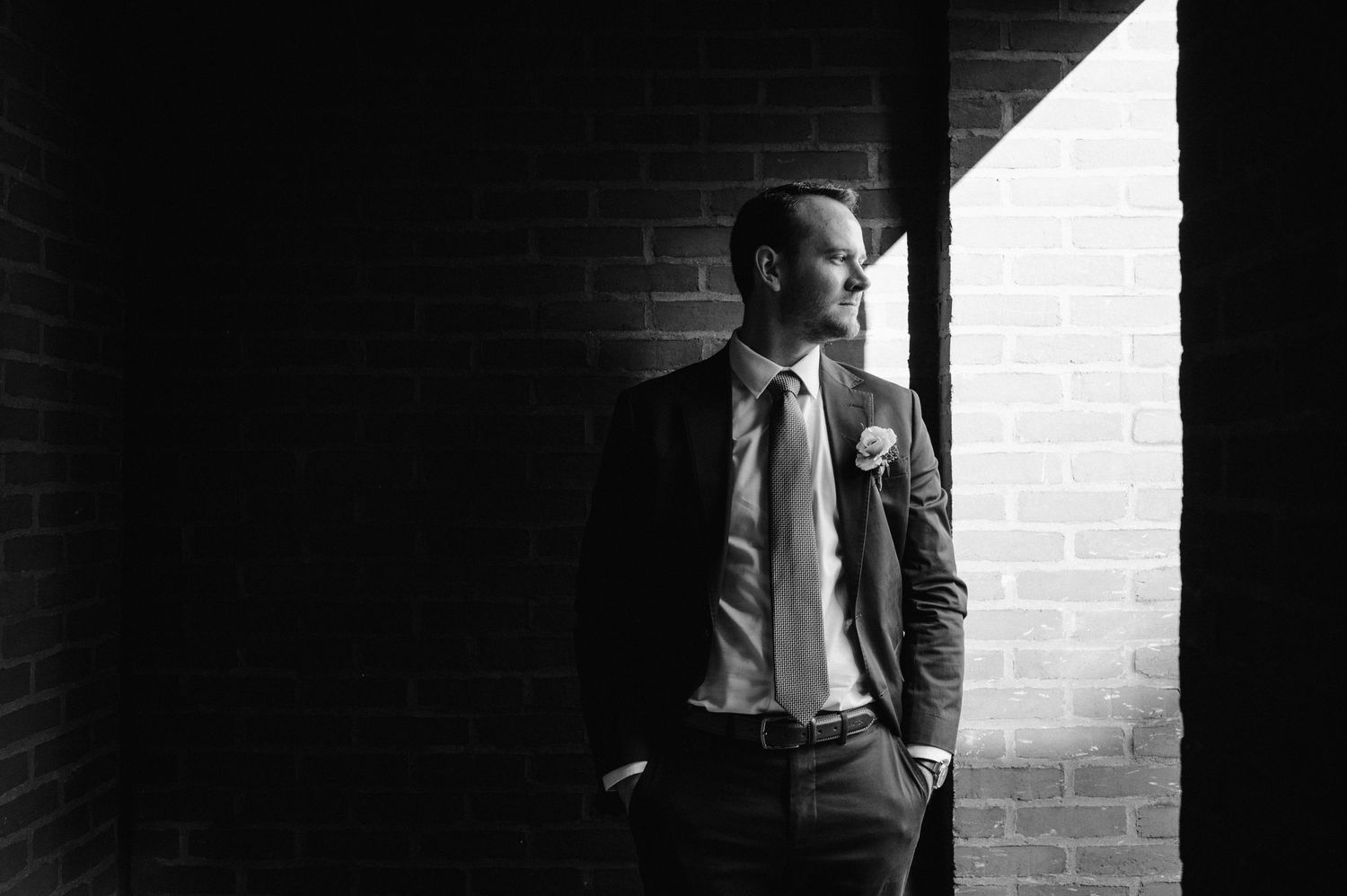 Black and white portrait of a suited person standing in dramatic window light against brick wall.