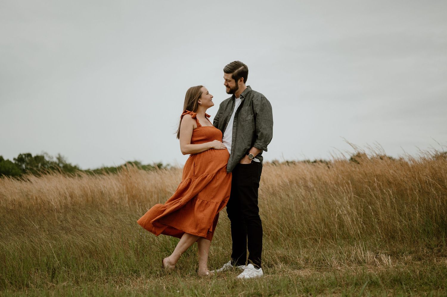 Romantic maternity photo of couple standing in tall grass during golden hour.