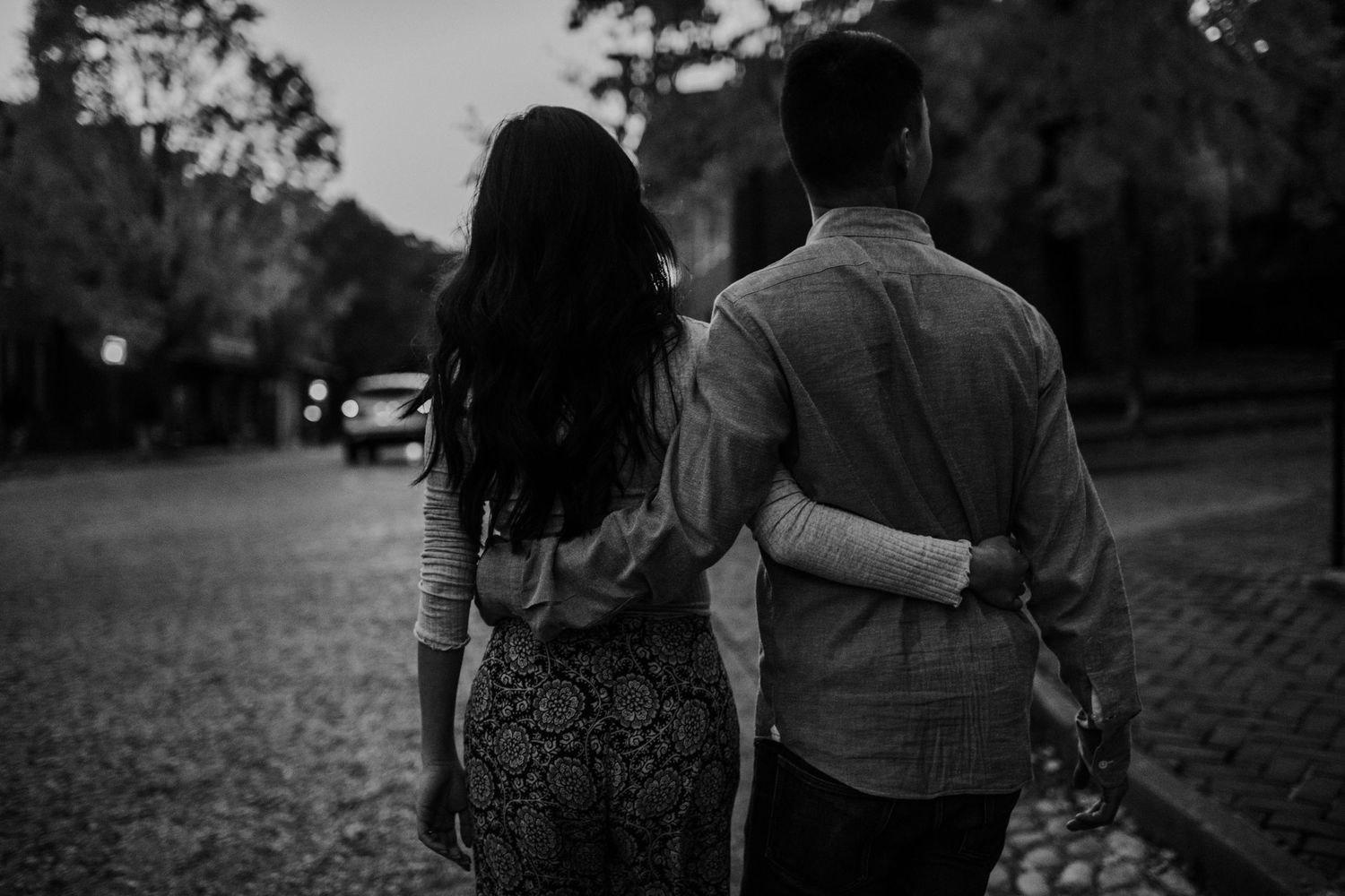 A couple walking together at dusk on a tree-lined street, viewed from behind.