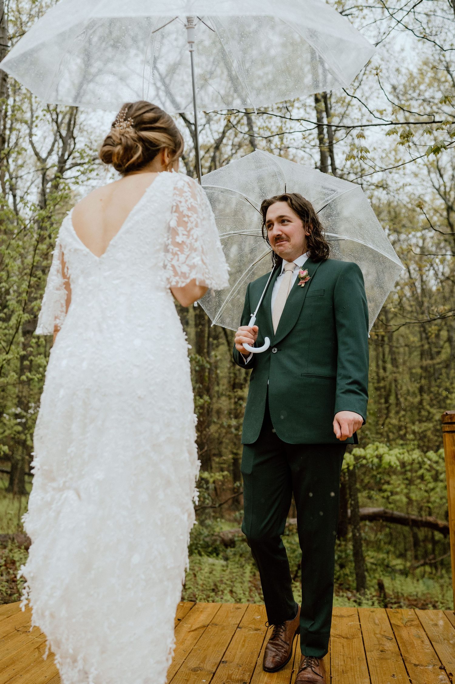 Wedding couple with umbrellas share emotional moment on wooden deck in forest setting.