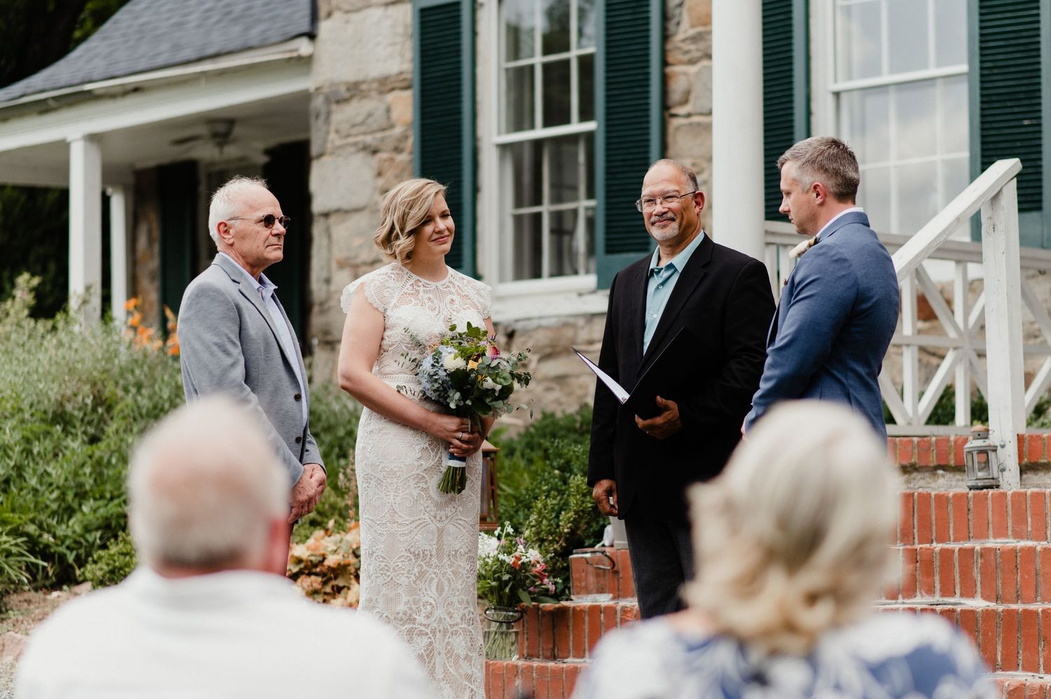 Outdoor wedding ceremony on brick steps of historic stone house with garden backdrop.