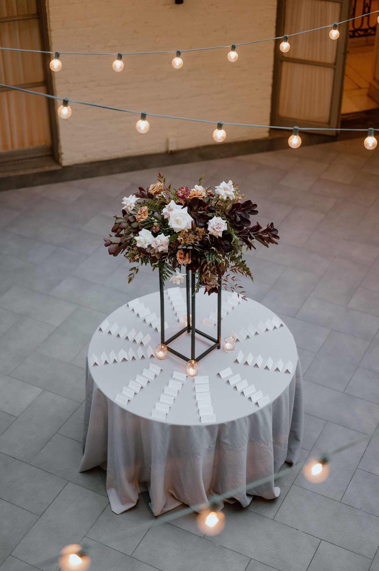 Wedding place card table with elegant floral centerpiece and string lights creating romantic ambiance.