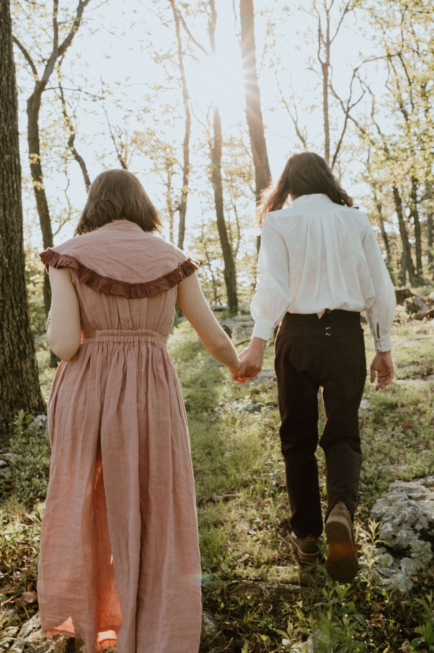 Couple holding hands walking through a sunlit forest path.