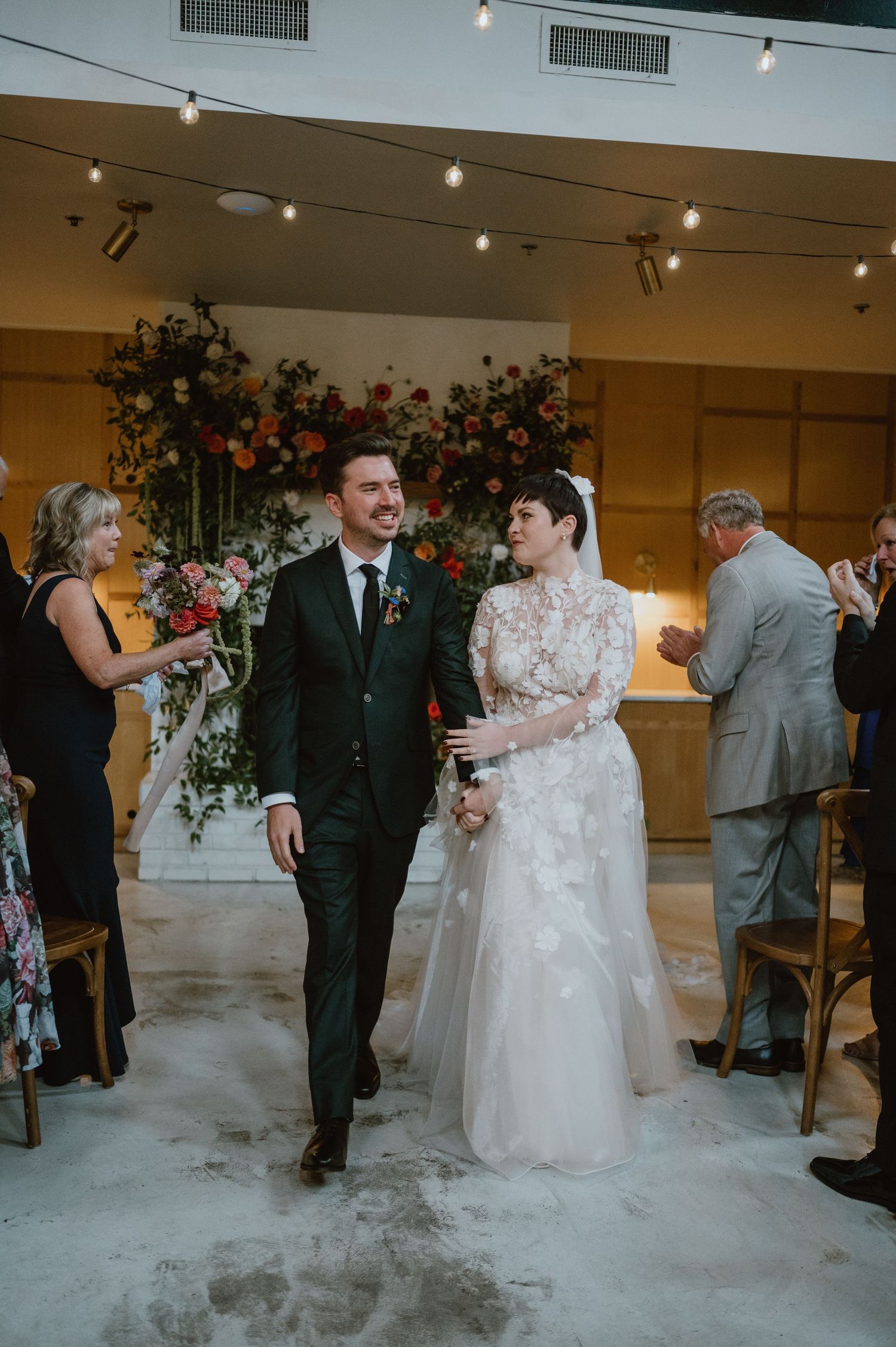 A wedding couple walks down the aisle together surrounded by floral arrangements and string lights in a modern venue.