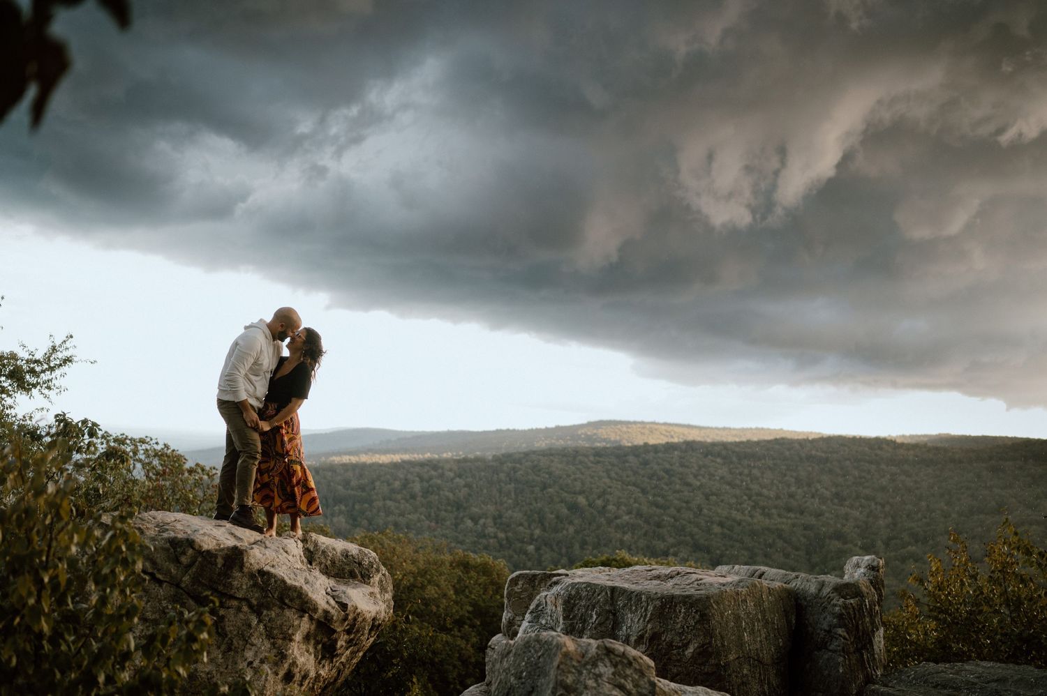 A romantic couple stands silhouetted against storm clouds on a rocky mountain ledge.