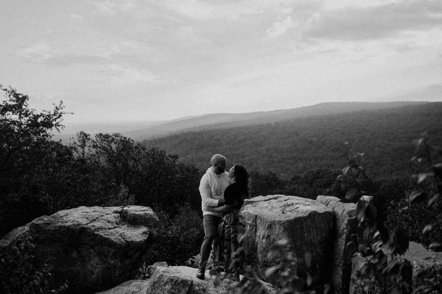 A silhouetted couple standing close together on rocky mountain overlook at sunset.