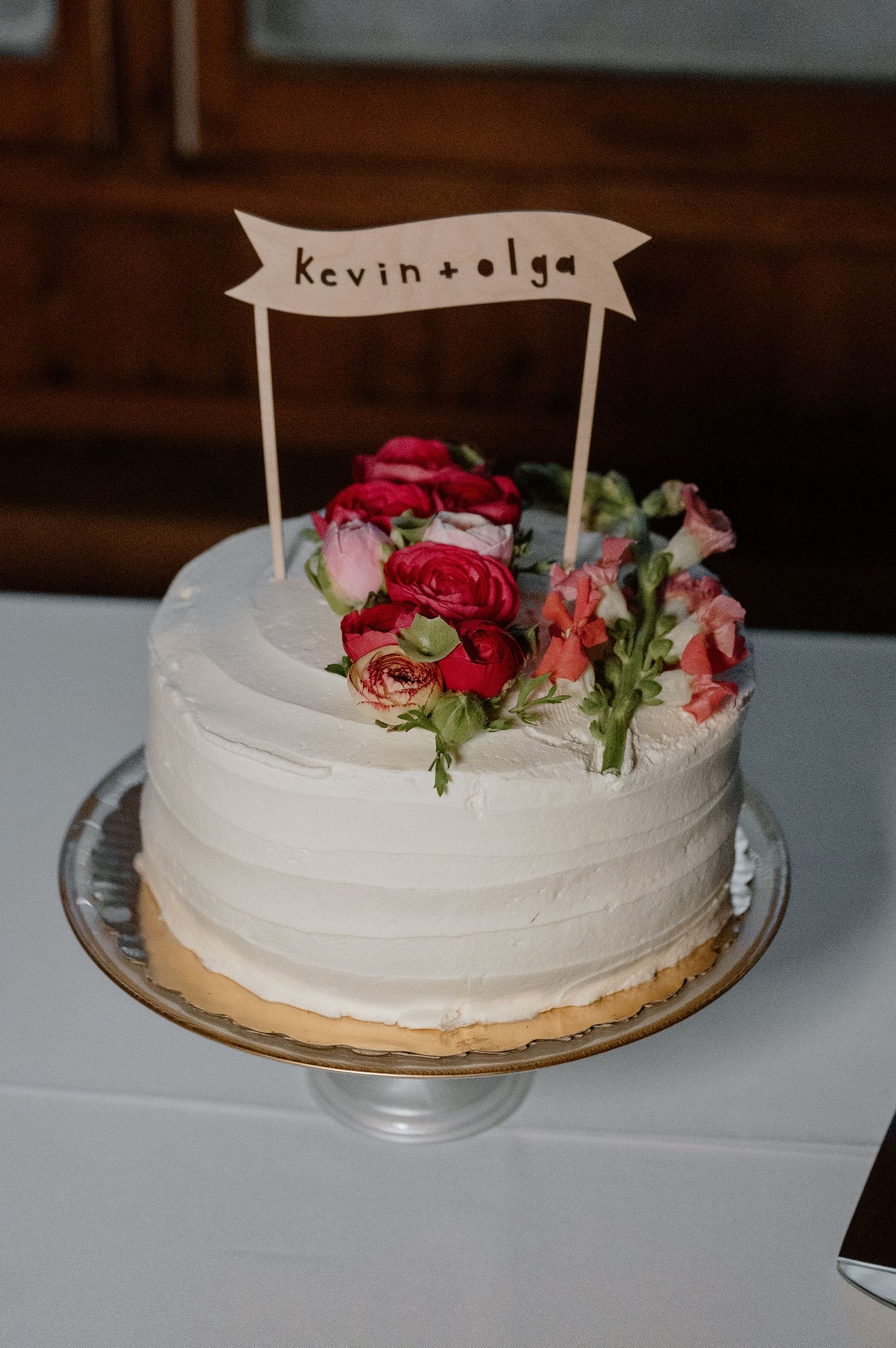 White wedding cake decorated with pink roses and a banner topper on a glass stand.