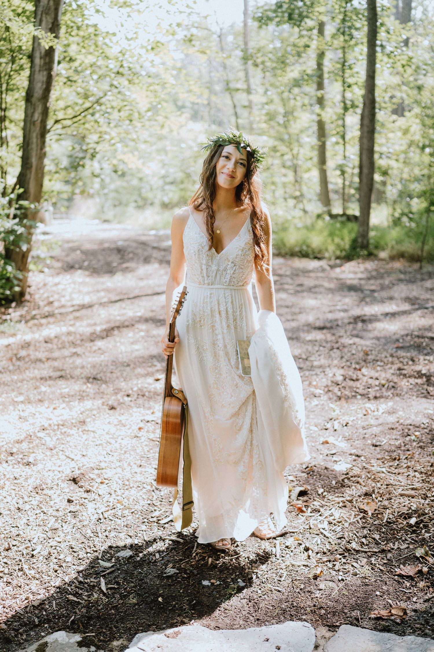 Person in flowing white dress holds guitar while standing on forest path in sunlit woods.