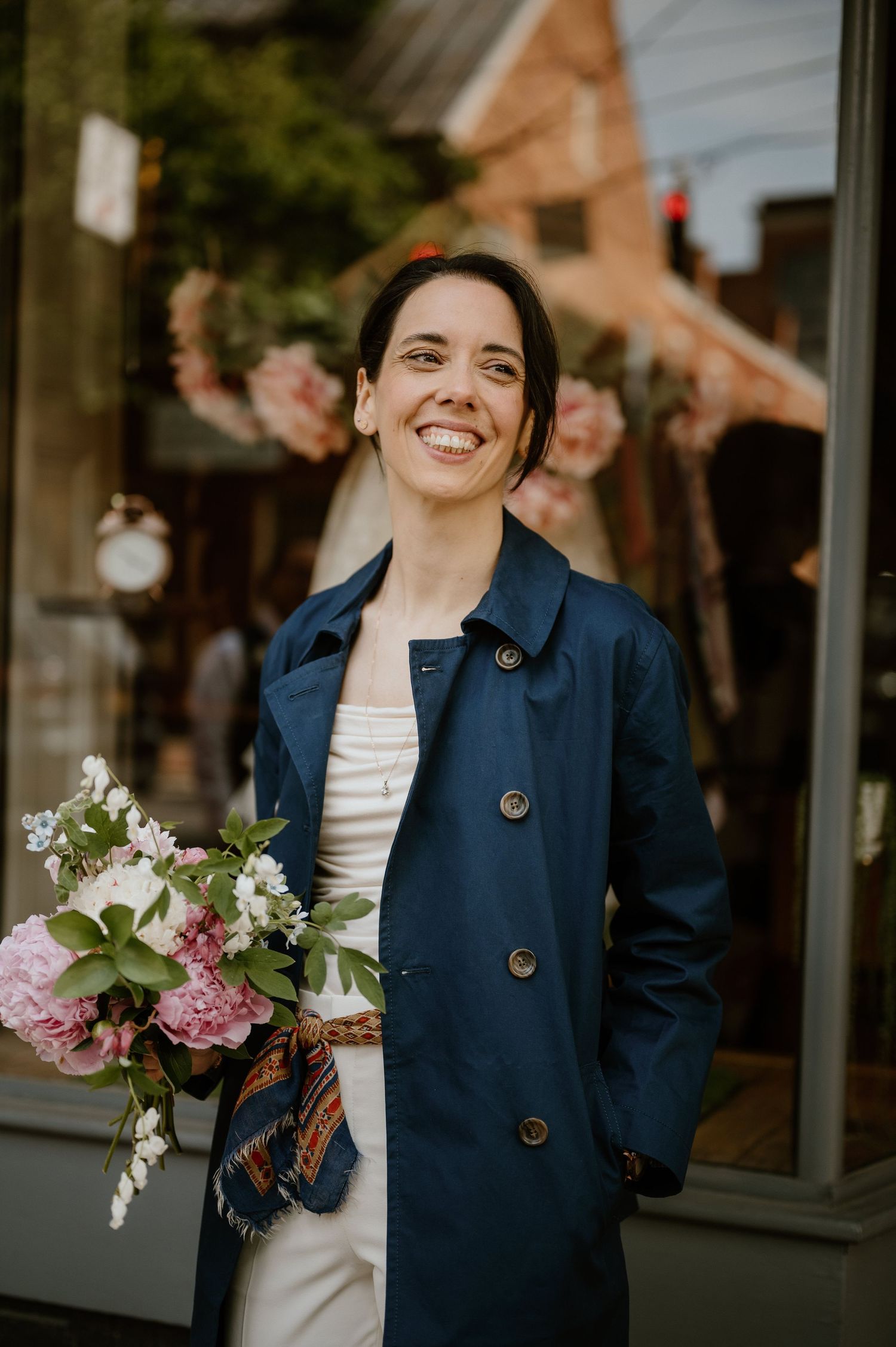A person wearing a navy coat holds a bouquet of pink and white flowers while smiling outside a shop.