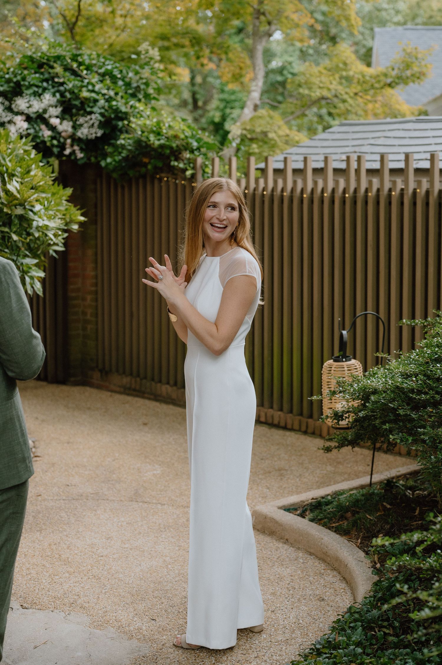 Person in white dress poses cheerfully in garden path with wooden fence backdrop.