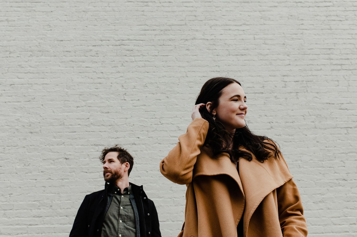A smiling woman in a tan coat and a bearded man in a dark jacket stand before a white brick wall.
