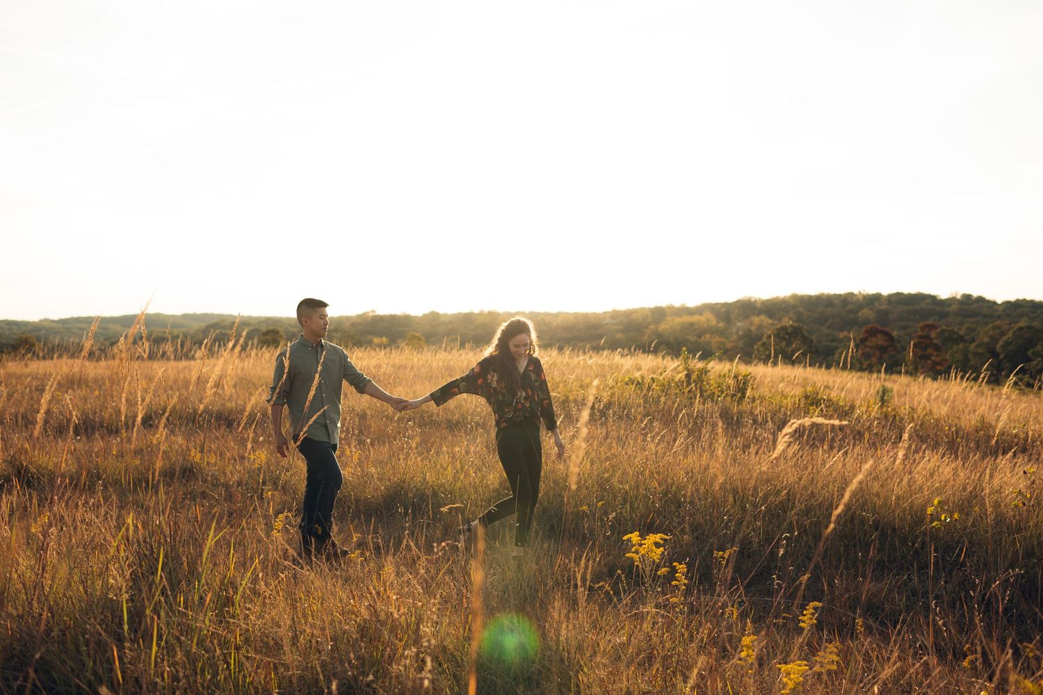 Couple holding hands walking through golden sunlit field at sunset in a romantic outdoor setting.