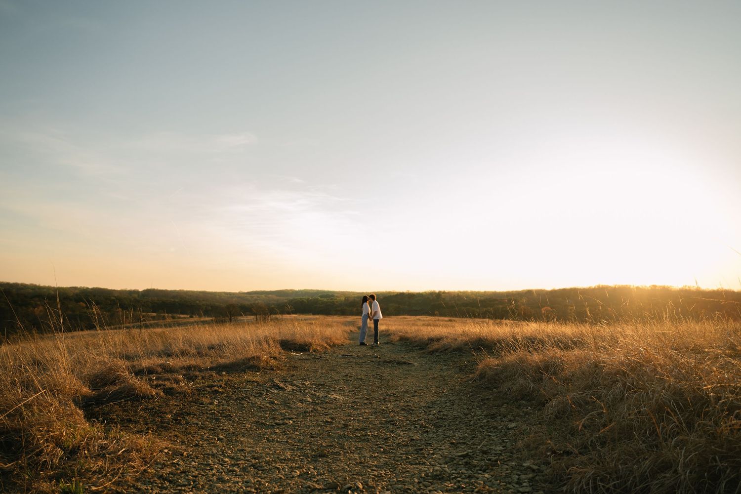 A couple walks along a dirt path through golden fields at sunset in a serene rural landscape.
