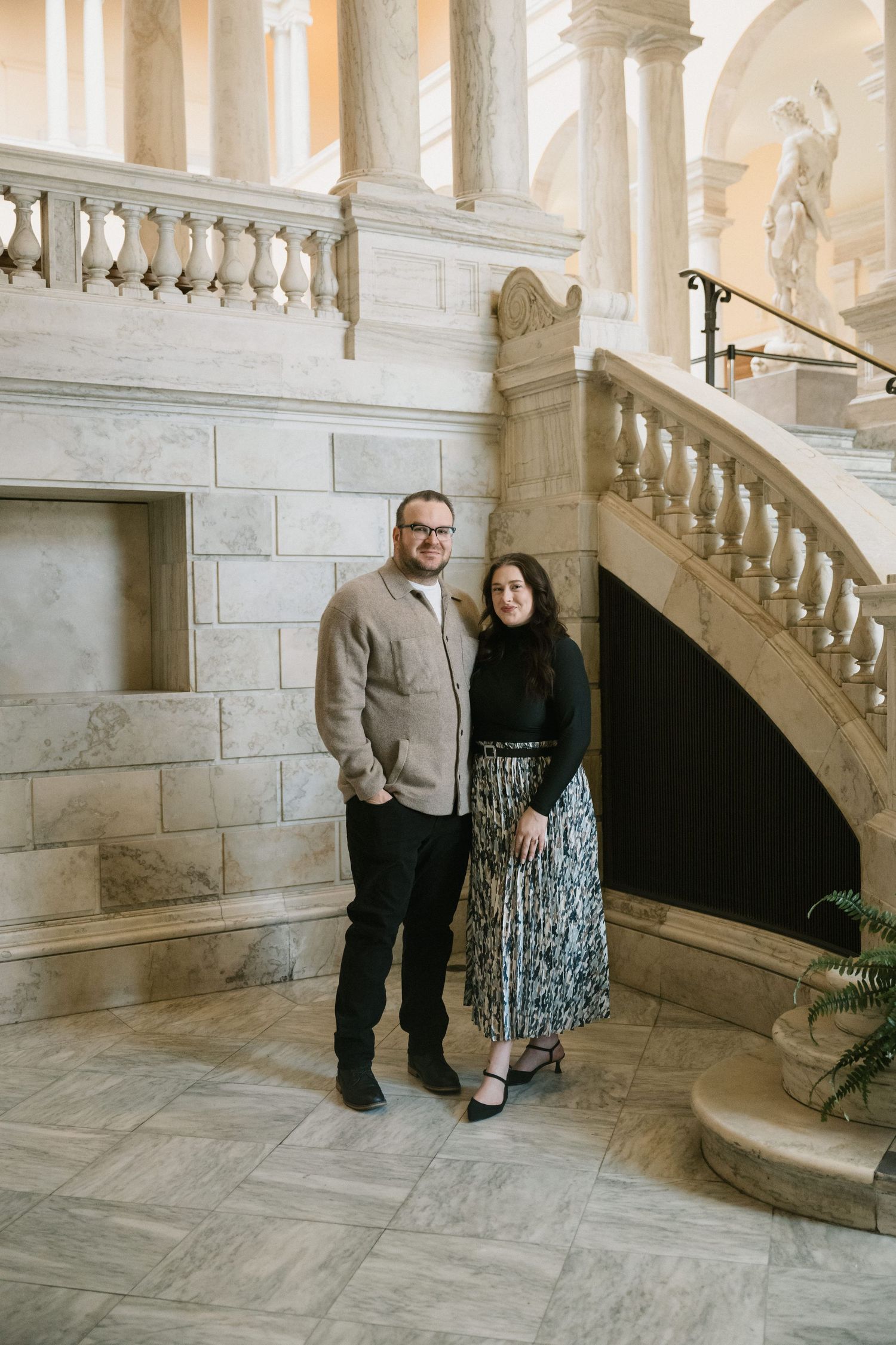 Couple posing together on marble staircase in elegant building interior, man in beige jacket and woman in floral skirt.