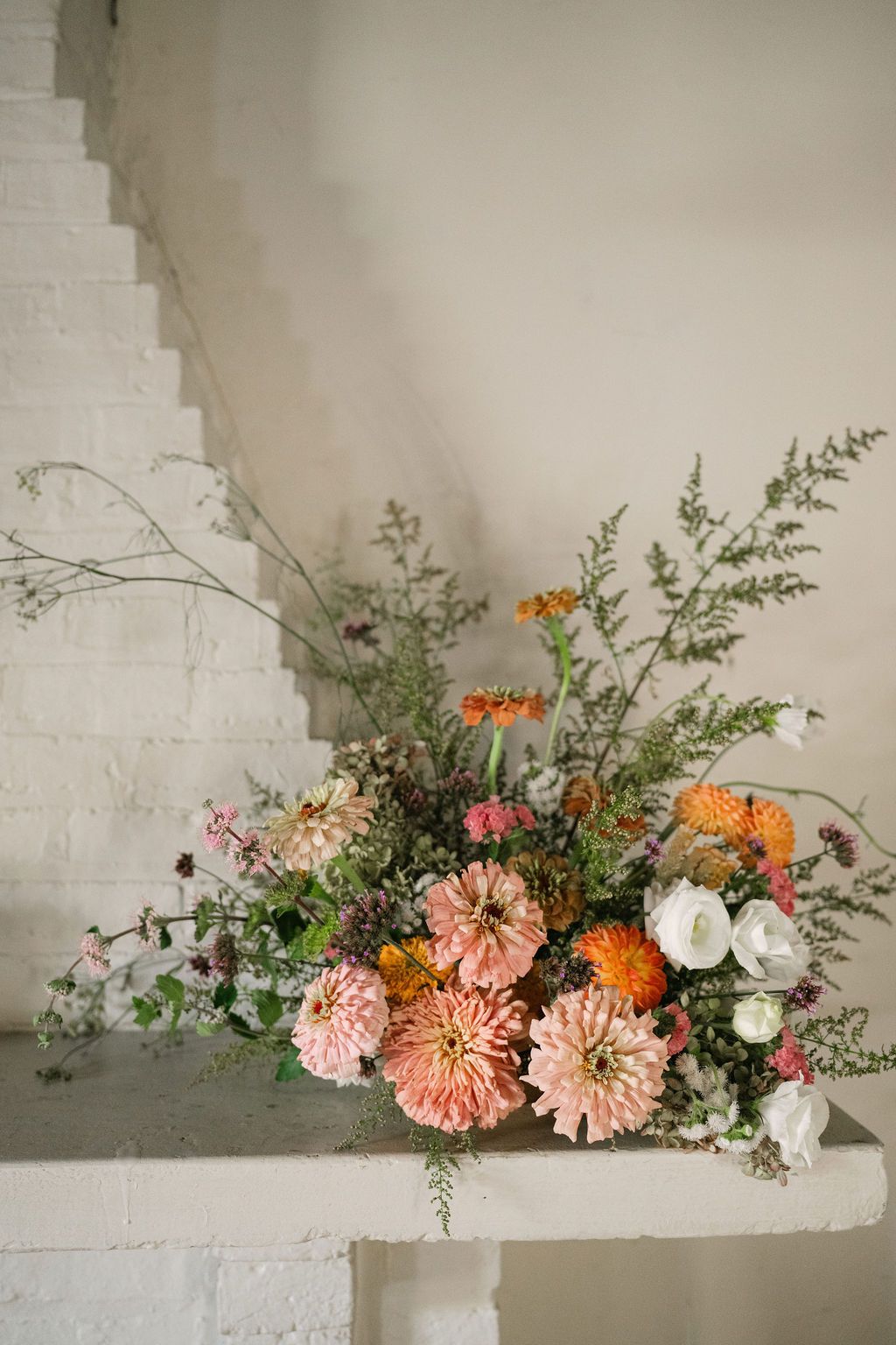 A rustic floral arrangement with coral dahlias, white flowers and greenery displayed on white stairs.