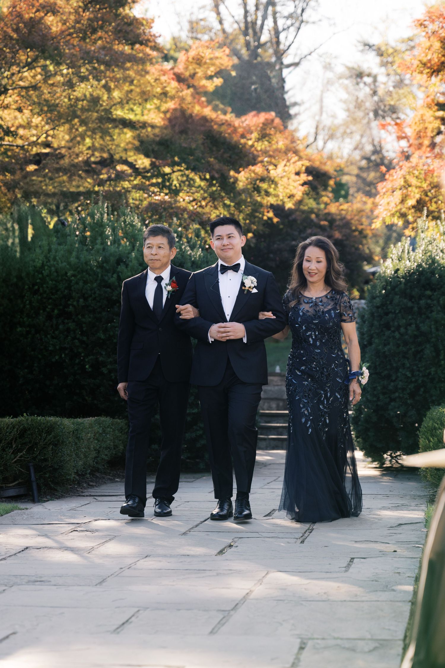 Three people in formal attire stand on a stone path surrounded by autumn foliage at a wedding.