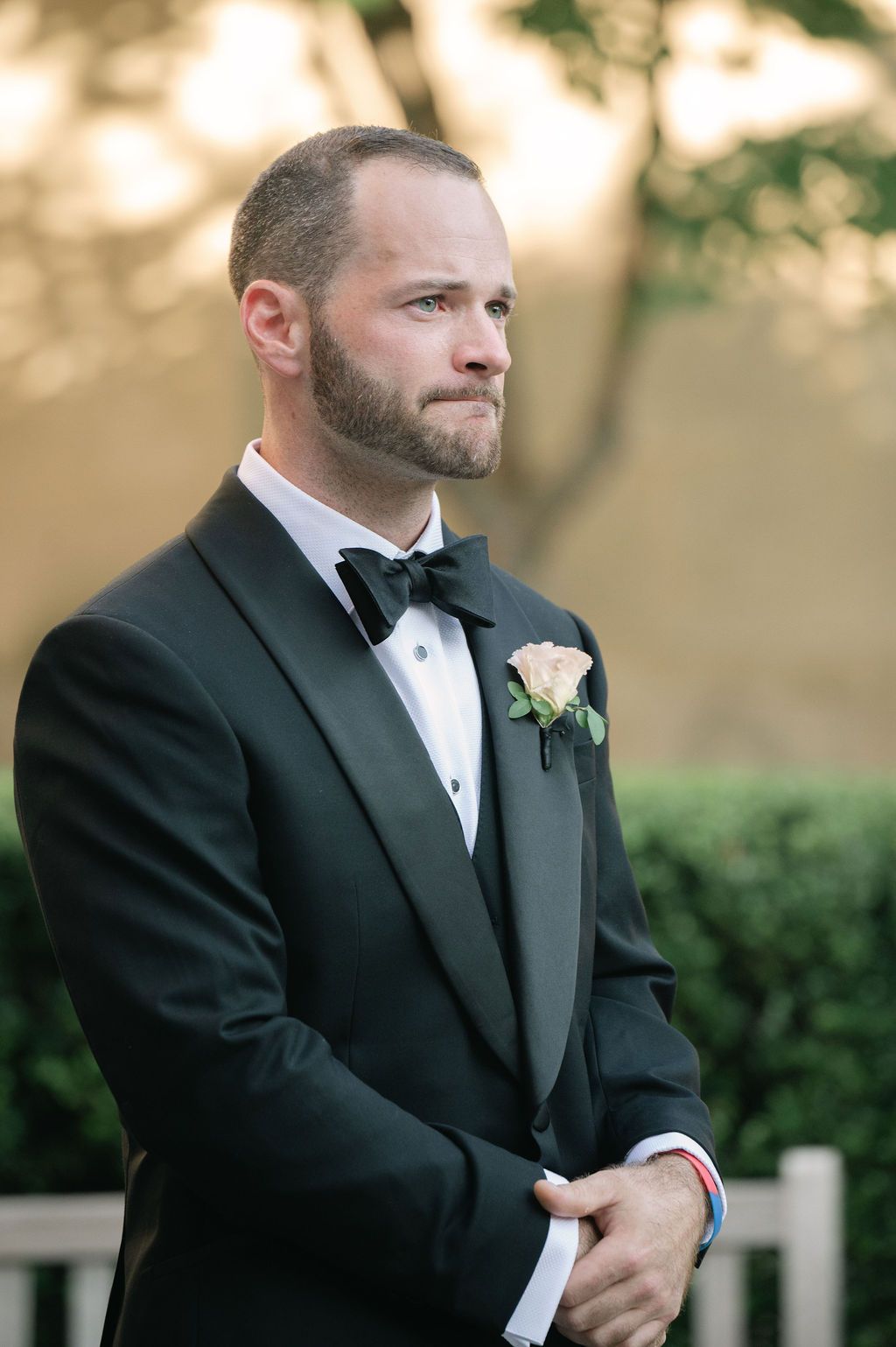 A groom in a classic black tuxedo and bow tie with a white boutonniere poses outdoors during golden hour.