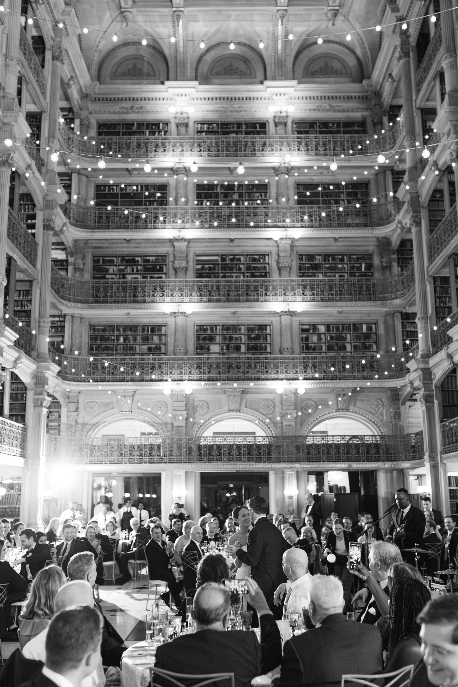 Black and white photo of an elegant library wedding reception with string lights and guests dining among multi-story bookshelves.