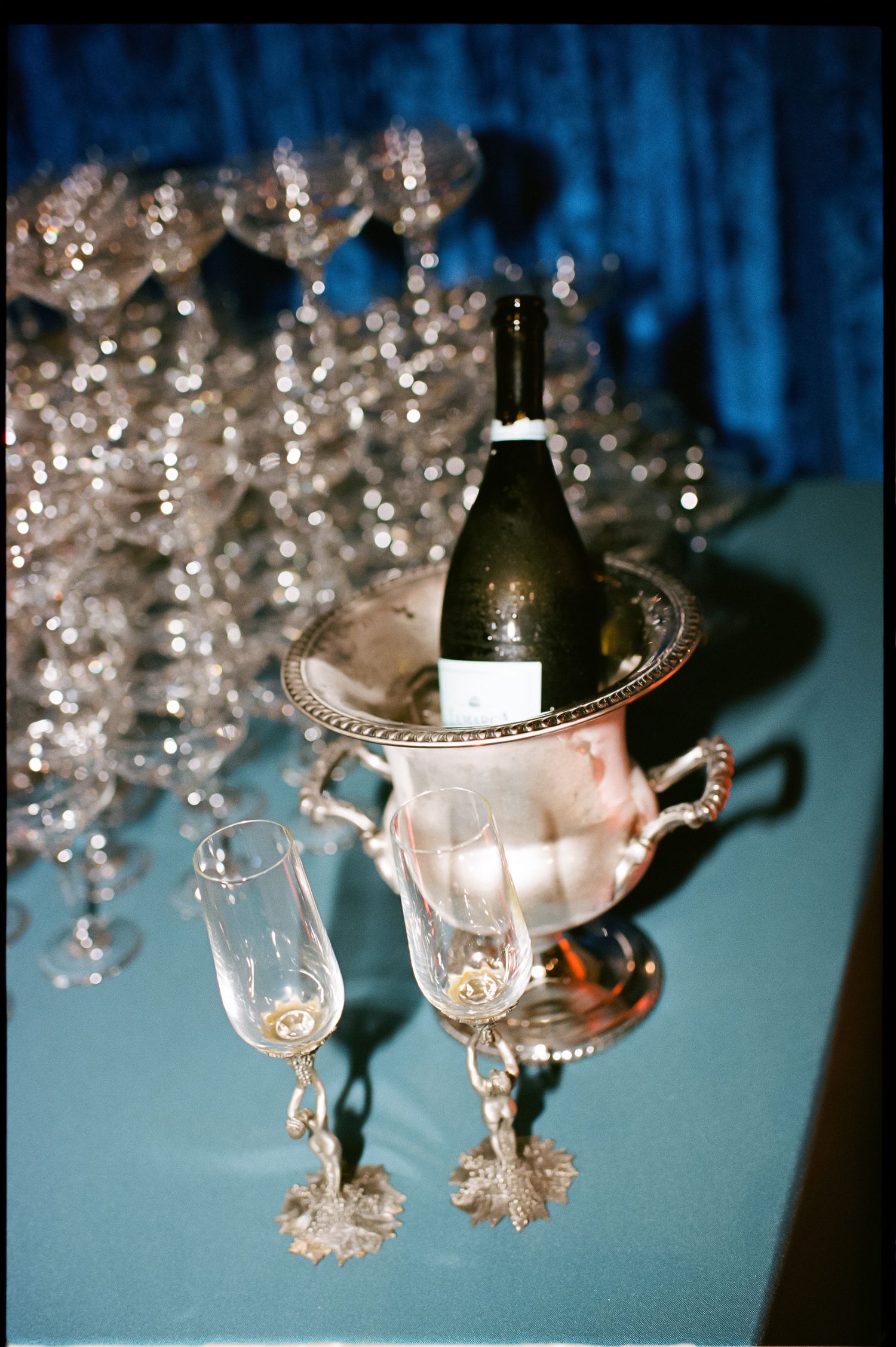 Elegant champagne bottle in silver ice bucket with crystal flutes and glassware on blue tablecloth at formal event.