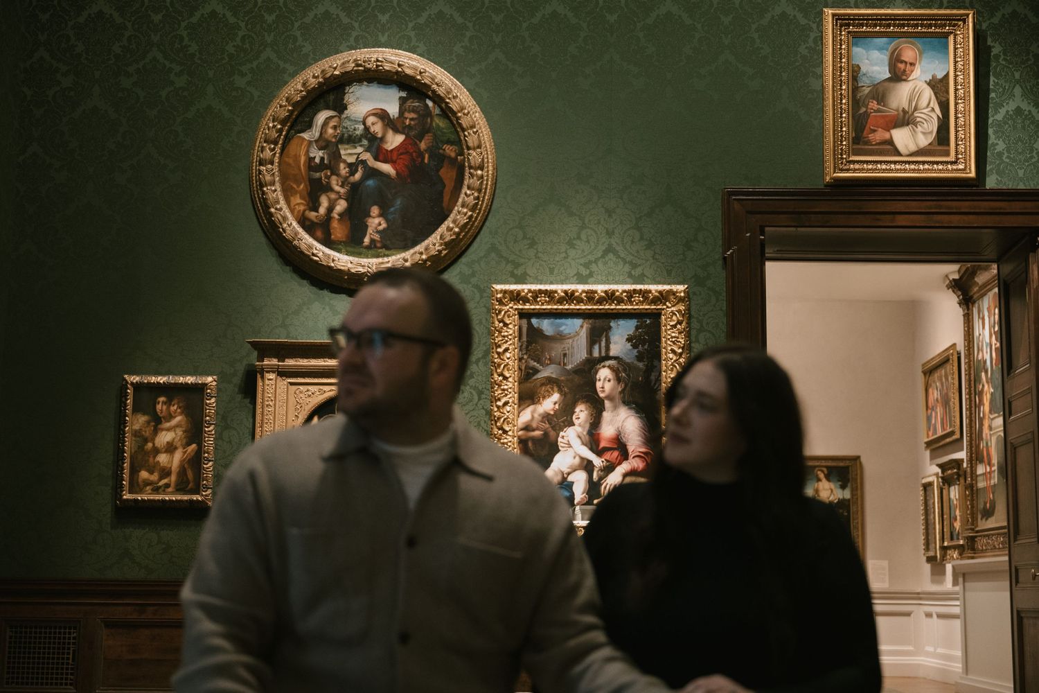 Two visitors stand before ornate Renaissance paintings in a green-walled art museum gallery.