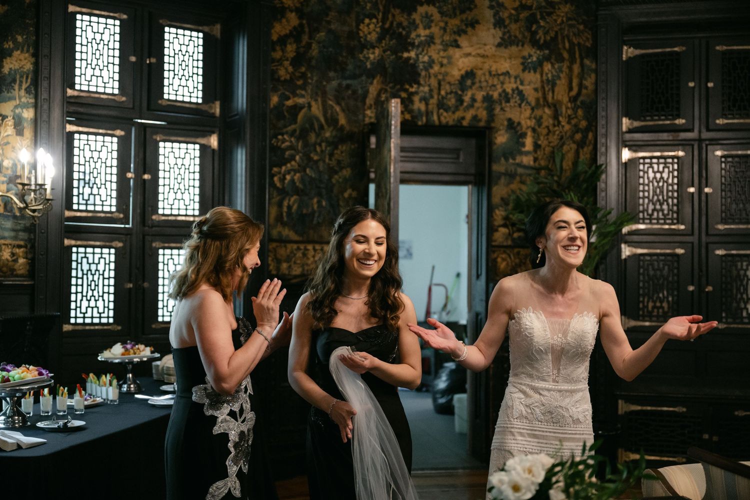 Three wedding guests share a joyful moment in a formal room with patterned wallpaper and decorative windows.