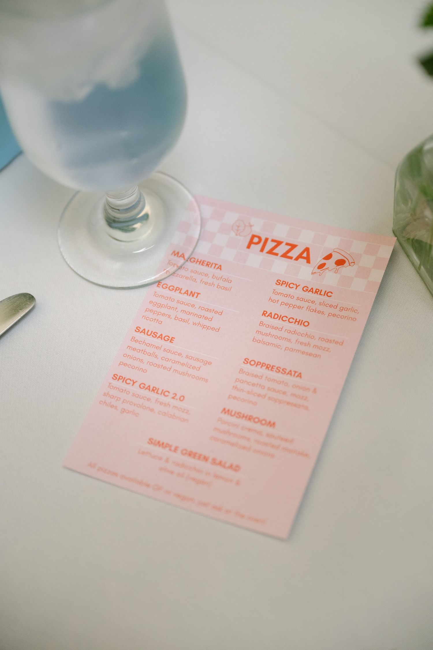 A pink menu card for pizza and appetizers placed on a white tablecloth at a formal dining setting.