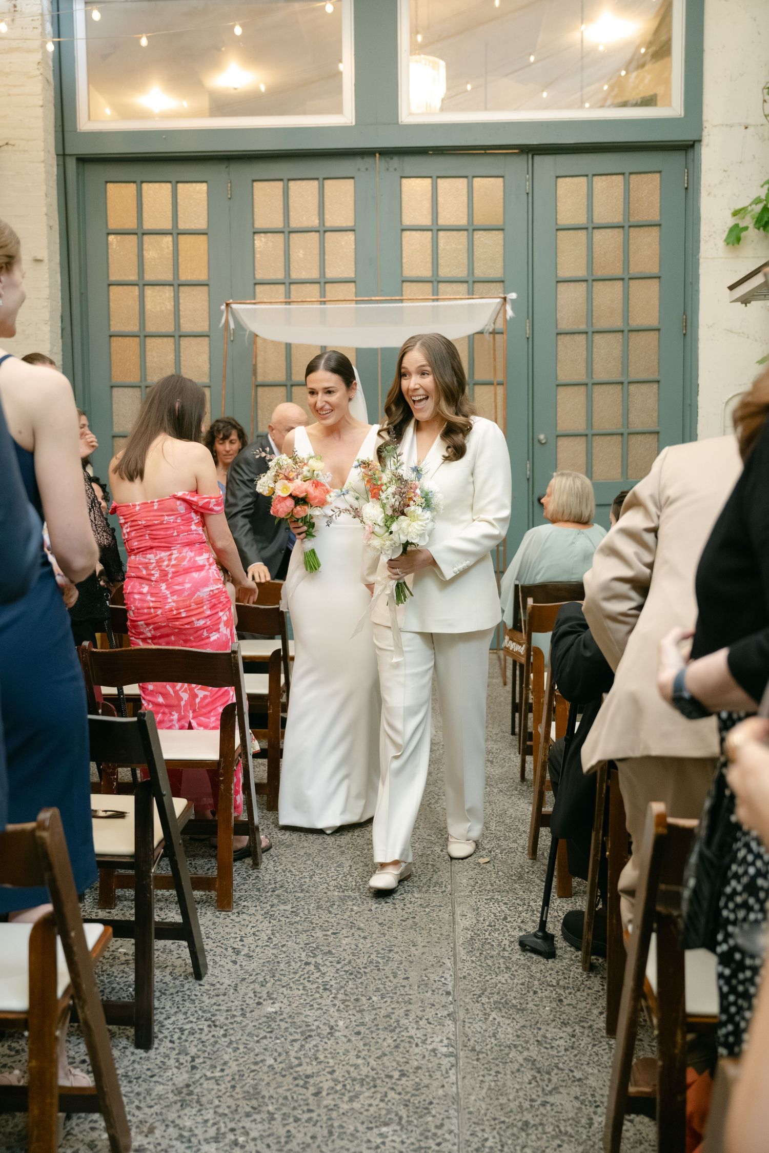 Wedding guests mingling and dancing at an indoor venue with string lights and wooden chairs at dusk.