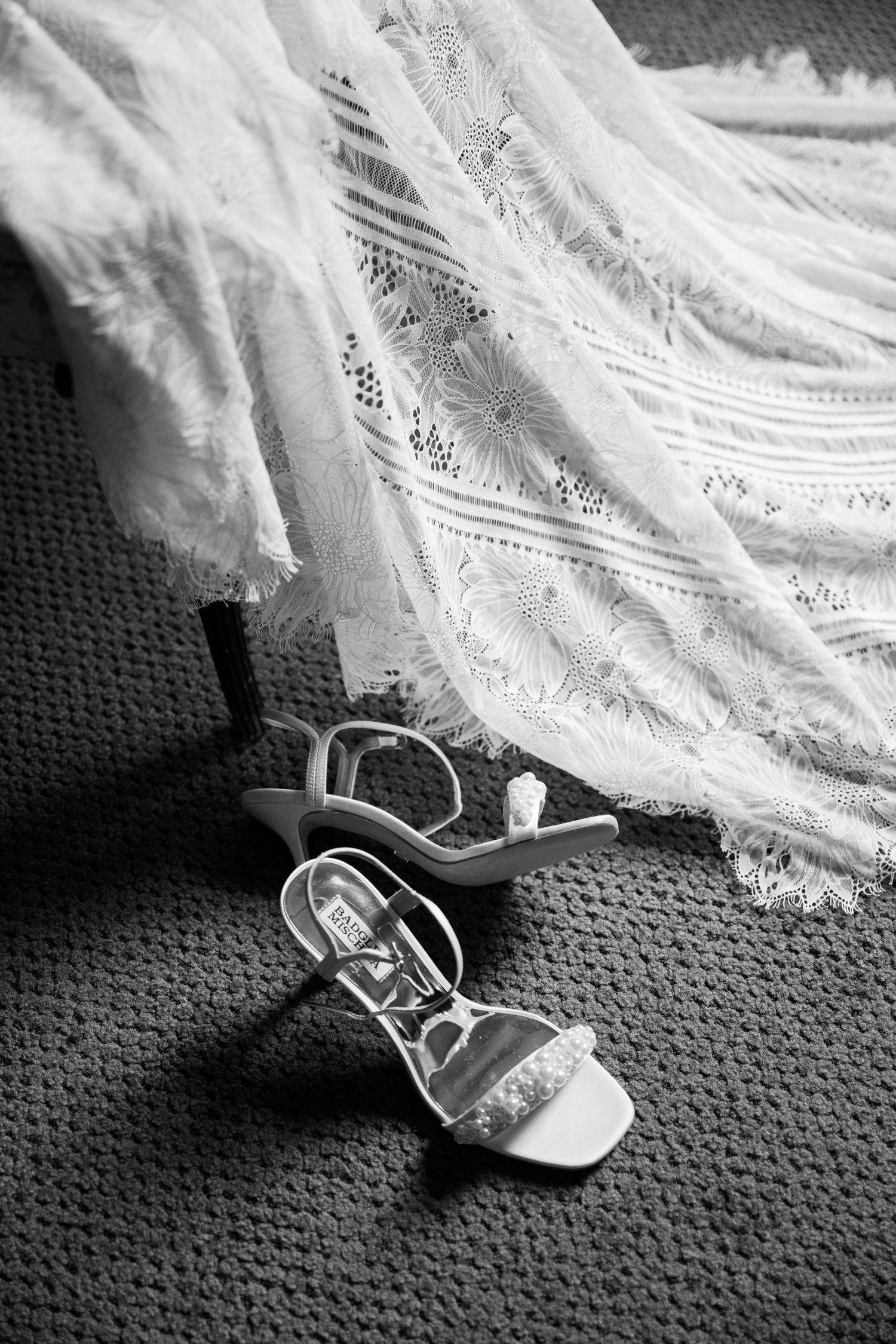 A delicate white lace wedding dress fabric drapes over a dark shoe on textured carpet in black and white.